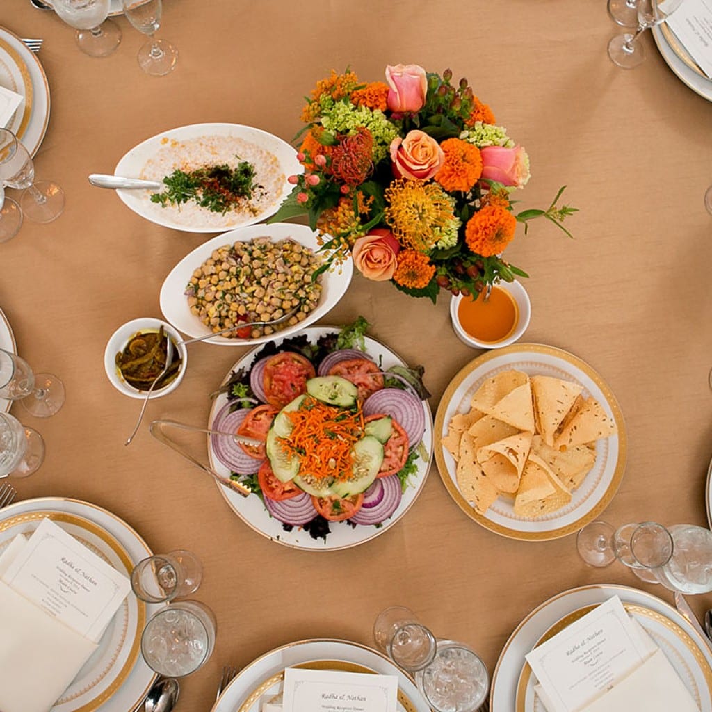 Family-style Indian dishes photographed from above on a set wedding reception table with printed menu cards.