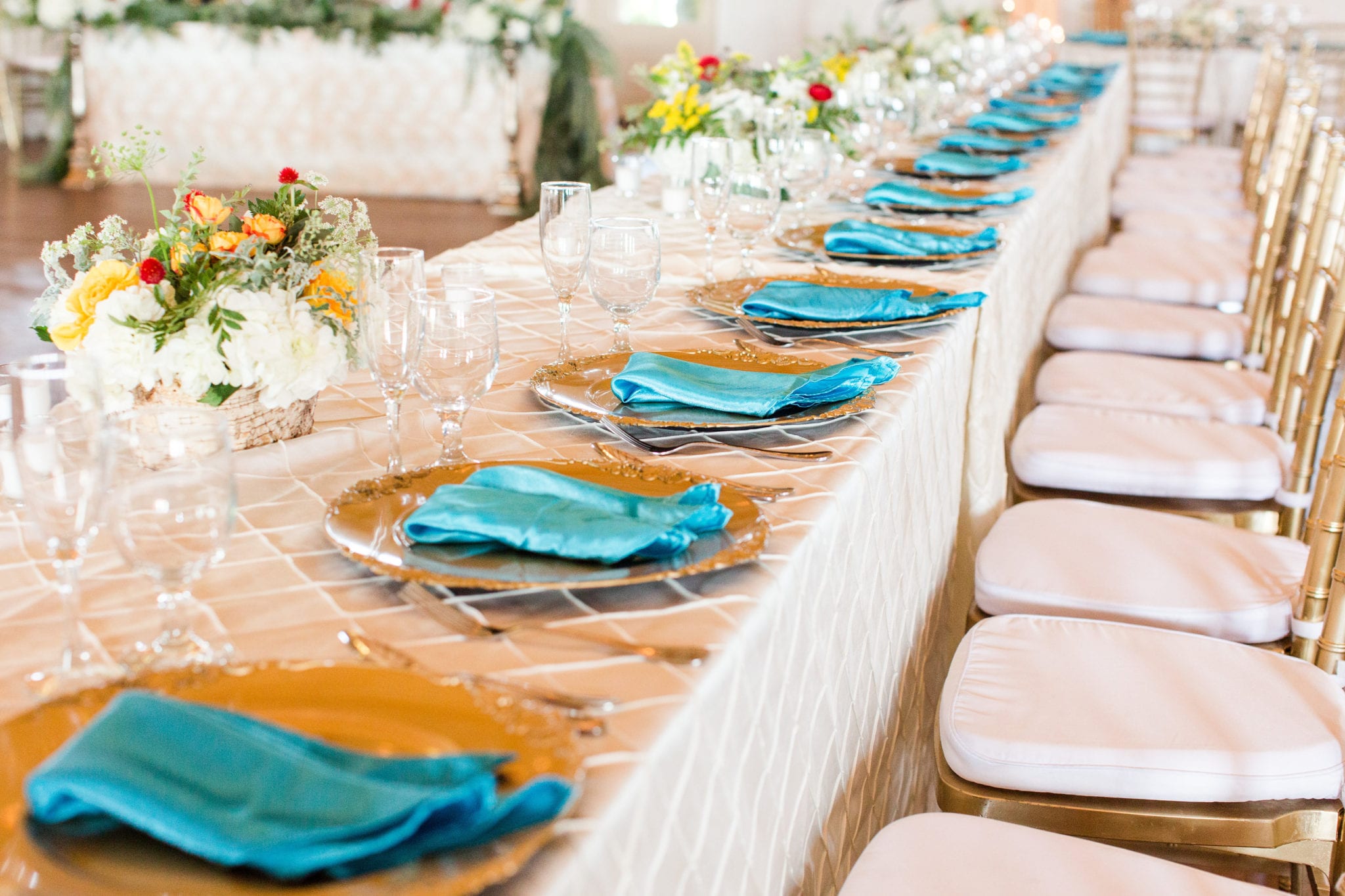 A long banquet table set for an IndAroma wedding reception — gold chargers, teal napkins and low floral centrepieces receding down the room.