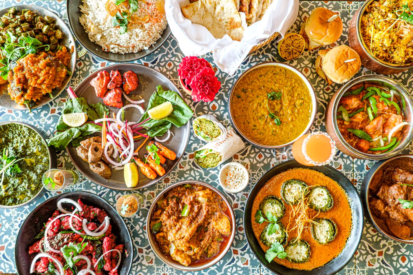 An overhead spread of IndAroma dishes on patterned tile — biryani, dal, palak paneer, kadhai chicken, tandoori chicken, a basket of naan, vada pav and chilli chicken.